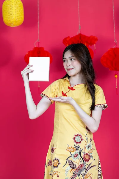 A young asian woman holding a white box for mockup while wearing simple yellow chinese dress, with some chinese ornament at the red background