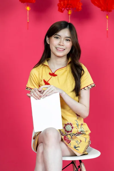 A young asian woman sitting and holding a white box for mockup while wearing simple yellow chinese dress, isolated on red background
