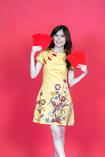 A beautiful asian woman sitting holding some hongbao red envelopes while wearing simple yellow chinese dress, isolated on red background