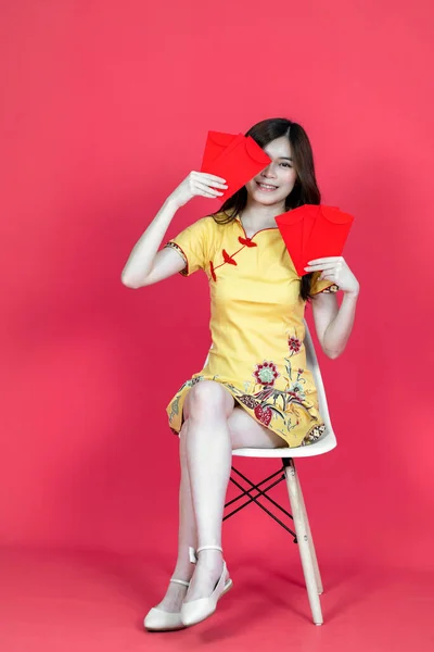 A beautiful asian woman sitting and holding some hongbao red envelopes while wearing simple yellow chinese dress, isolated on red background