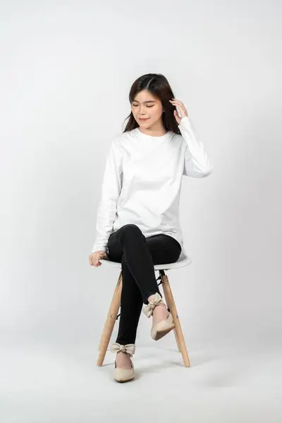 A young asian woman wearing a white blank long sleeve shirt doing a pose while sitting, isolated on the white background