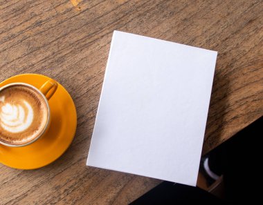 A white colored hardcover book with a wooden textured table as the background and a coffee near it, hardcover mockup template image