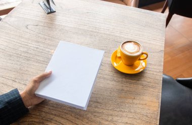 A hand holding a hardcover book with a wooden textured table as the background and a coffee near it, hardcover mockup template image