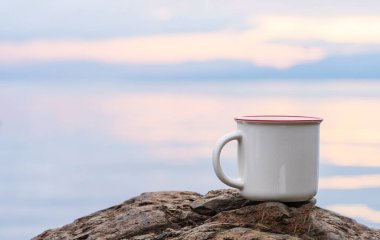 An enamel mug standing still on the top of a rock while showing the out of focus scenery at the background, enamel mug mockup image