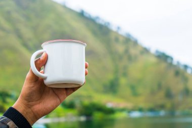 A hand holding an enamel mug while showing the out of focus scenery at the background, enamel mug mockup image