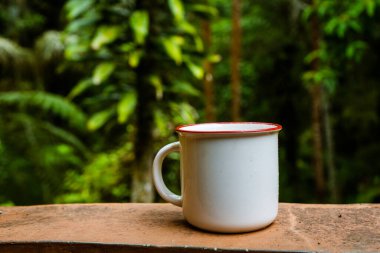 An enamel mug standing on the top of an orange fence with out of focus trees background, enamel mug mockup image