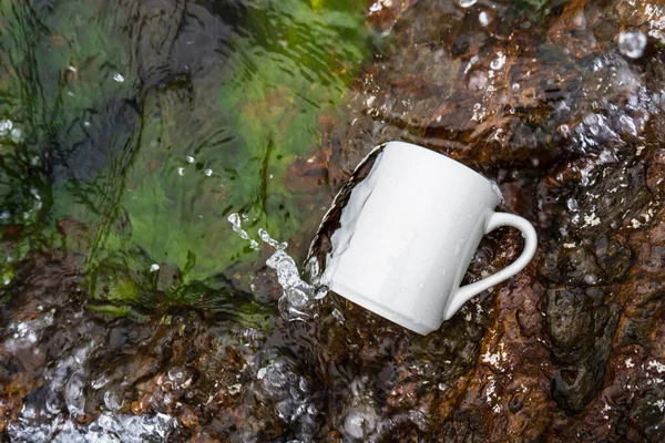 Coffee mug laying out on the top of some rocks that covered with moss while the water keep splashing over it, coffee mug mockup image