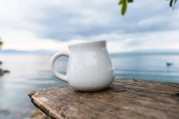 A cream mug stand still on the top of a piece of wood with an out of focus pond and sky as the background, cream mug mockup image