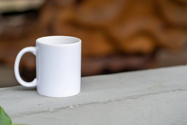 A blank white tea mug standing on the top of a cement fence with blurred out brown background, coffee mug mockup image