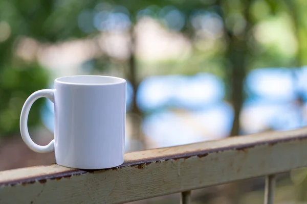 White blank coffee mug standing on the top of an iron fence with out of focus blue background, coffee mug mockup image