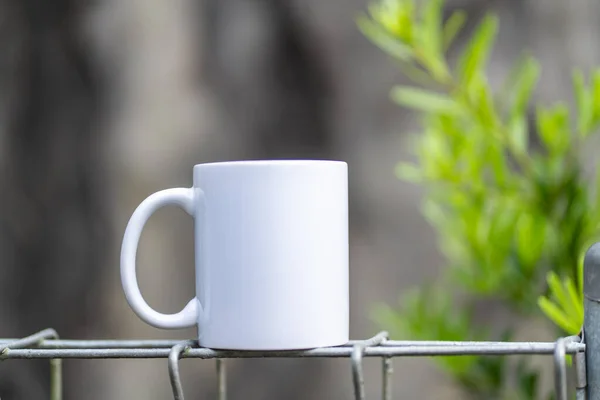 White blank tea mug standing on the top of a fence with a green plant as the blurred out background, coffee mug mockup image