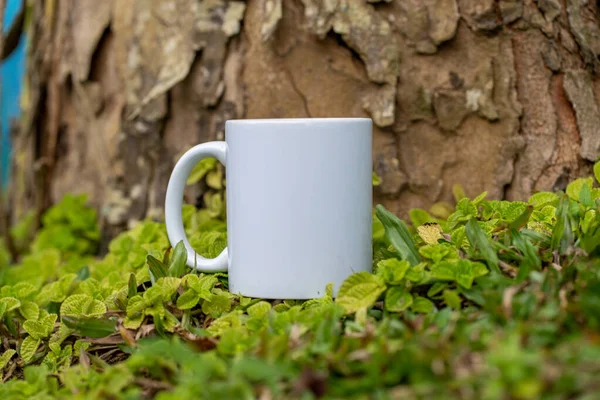 A blank white coffee mug standing out on the top of a grass with some green plants around it, coffee mug mockup image