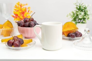 A white blank coffee mug on the top of a rounded mat with the fresh fruits arranged around it, coffee mug mockup image