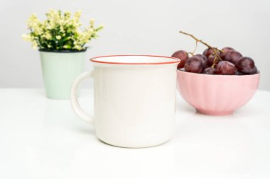 A white blank enamel mug on the top of a white table with the grapes in a bowl placed behind it, enamel mug mockup image