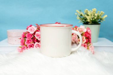 A white blank enamel mug standing out on top of a fluffy white mat with some flower behind it, enamel mug mockup image