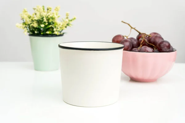 A white blank enamel mug on the top of a white table with the grapes in a bowl placed behind it, enamel mug mockup image
