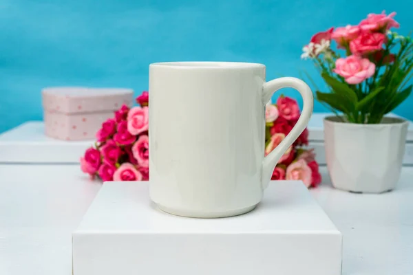 A white blank coffee mug standing out on top of a white box with some flower behind it, coffee mug mockup image