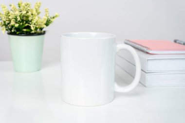 A white blank coffee mug on the top of a white table with some books decorated behind it, minimalistic looks, coffee mug mockup image
