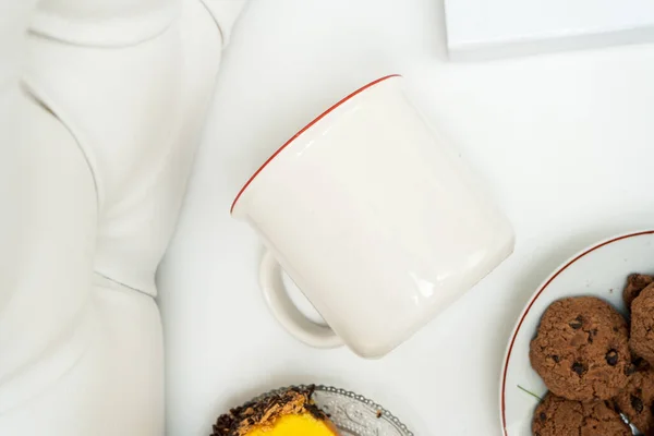 A white blank enamel mug laying out on the top of a white sofa with some snacks placed near it, enamel mug mockup image