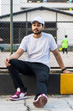 Genç sakallı adam beyaz gömlekli ve şapkalı, arkasında tenis kortu olan beyaz şapkalı ve gömlekli bir adam.