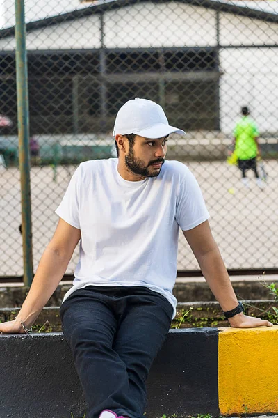 Entellektüel sakallı adam, bulanık tenis kortu geçmişi, beyaz boş şapka ve tişört modeli görüntüsü ile poz veriyor.