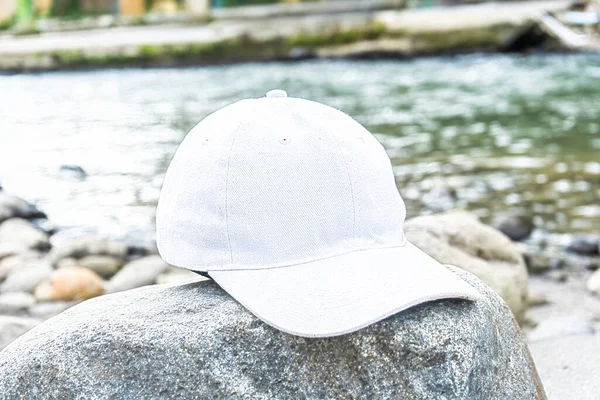 a white cap hat is artfully displayed above a rock, creating an elegant and stylish aesthetic near the river, white blank cap hat mockup image
