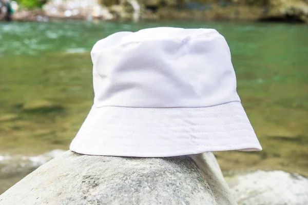 The pristine white bucket hat featured in this photo is tastefully positioned above a rock near the tranquil waters of the river, white blank bucket hat mockup image