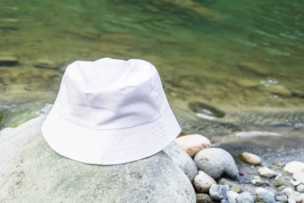 In this peaceful setting, a white bucket hat is elegantly displayed on a rock near the gently flowing river, white blank bucket hat mockup image