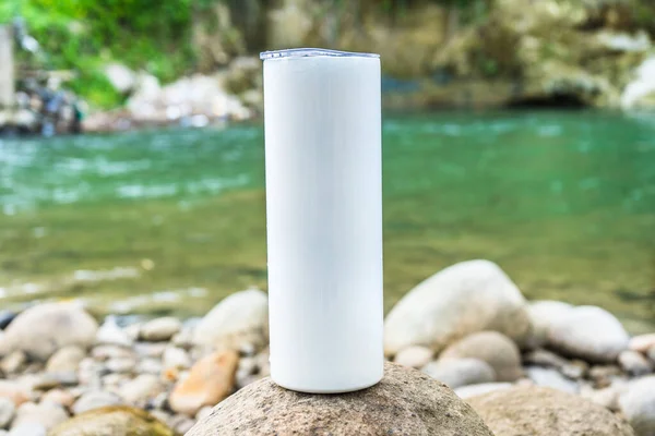 In this tranquil scene, a long tumbler is artfully displayed above a rock near the serene waters of the nearby river, adding a touch of sophistication to the setting, white blank long tumbler mockup image