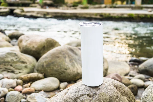 stock image The clean and elegant design of this long tumbler is beautifully captured in this photo, positioned above a rock near the serene river, white blank long tumbler mockup image