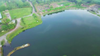 Yukarıdan çekilen manzara, sakin bir köy, parıldayan bir gölet ve yemyeşil palmiye tarlaları içeriyor.