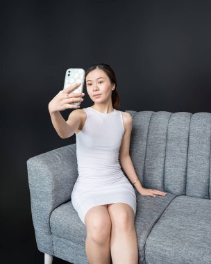 Göz alıcı Asyalı bir kadının portresi rahat bir kanepede, büyüleyici bir bakışla selfie çekiyor. Gömlek modeli bir resim.