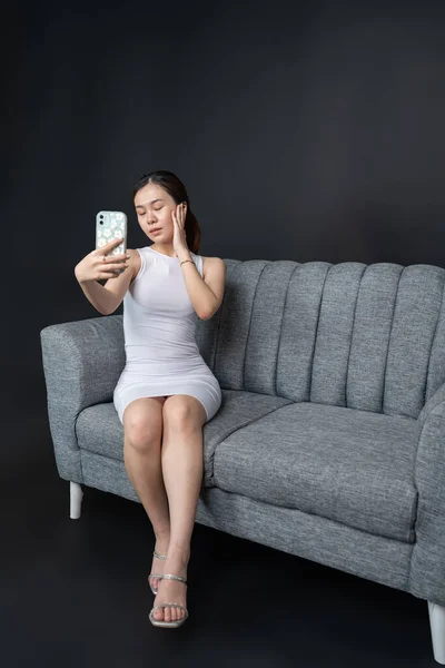 Indoor portrait of an alluring Asian woman capturing her beauty in a selfie on a luxurious sofa, shirt dress mockup template image