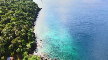 Bir ada ile mavi deniz suyu arasındaki kesişmeyi gösteren hava görüntülerinin sonunda açık bir gökyüzü olan bir dron ile çekilmiş.