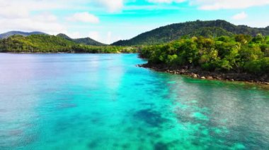 Mavi deniz suyunun görüntüsü ve adanın berrak gökyüzü alçak irtifalı bir dron tarafından sağdan ve önden kuşatılmış.
