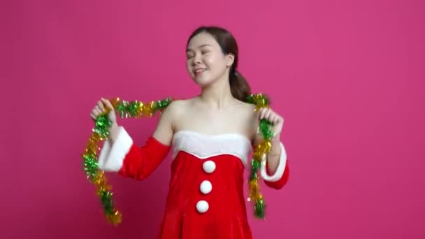 一个亚洲女人穿着以圣诞节为主题的衣服快乐地跳舞 同时背带着粉色背景的圣诞装饰品的形象 — 图库视频影像