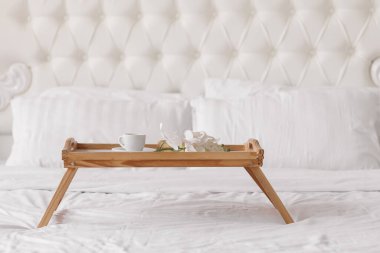 Romantic morning. Wooden coffee table with flowers on bed with plaid, coffee cup, flowers and donut. Interior white tones.