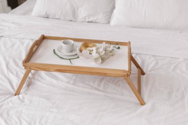 Romantic morning. Wooden coffee table with flowers on bed with plaid, coffee cup, flowers and donut. Interior white tones.