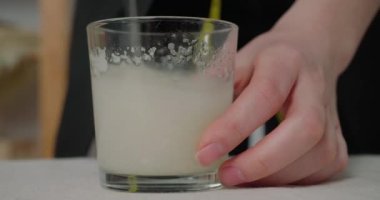 Woman makes glue mixing liquid with spoon in glass standing at table. Milliner makes mixture to smear on handmade hat in workshop closeup