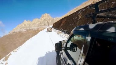 Cars drive on dangerous snowy road in giant Caucasus mountains under clear blue sky. Extreme driving in highland on sunny winter day closeup
