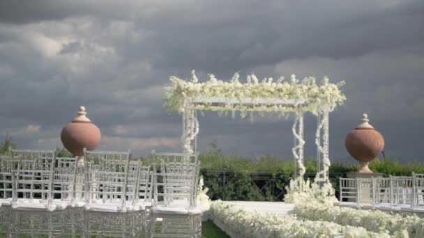 银道上装饰着白花的婚礼拱门 在多云的天空下 有白色椅子和草坪上花卉装饰的小巷 — 图库视频影像