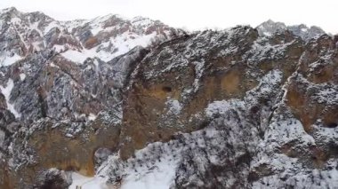 Caucasus mountain range with rocky peaks in white snow against cloudy sky. Bare trees grow on steep slopes of highland in winter aerial view