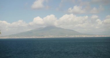 Güneşli Sorrento yakınlarında uzak sisli volkan Vesuvius 'a karşı masmavi deniz yüzeyi. Kümülüs bulutları güney İtalya 'daki devasa volkanik tepeyi kaplar.