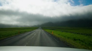 Araç kırsal yol boyunca gri bulutların altında ilerliyor. Hava koşulları karanlık yaratıyor ve sürüş deneyimini karmaşıklaştırıyor
