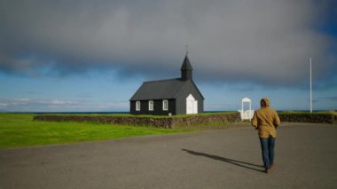 Ceketli adam İzlanda 'da yeşil çimlerle çevrili Budakirkja Kilisesi' ne yürüyor. Gri bulutlar gökyüzünü yayarak mistik atmosfer yaratır