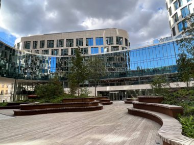 Kavisli cam cepheleri ve peyzajlı açık hava oturma alanlarının yer aldığı modern mimari Beobank Merkez Binası. Sahne bulutların yansımalarını yakalıyor ve sakin bir şehir ortamı yaratıyor..