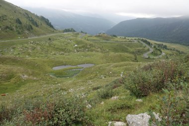 Bereketli yeşil tepeler ve bulutlu bir gökyüzünün altındaki küçük gölcüklerle çevrili dolambaçlı bir dağ yolunun manzarası..