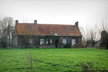 Aalter Flanders Belçika 1201 2024 terk edilmiş, eski, harap bir tuğla çiftlik evi. Hasarlı bir çatı ve kırık camları var. Etrafı çimlerle ve yapraksız ağaçlarla çevrili, bulutlu bir gökyüzünün altında..