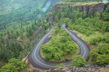 A Winding Road Through Forested Cliffs at Rowena Crest, Columbia river Gorge, Oregon curves through a lush green forested landscape with cliffs. clipart