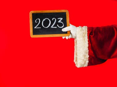 Noel Baba 'nın elinde tebeşirle yazılmış 2023 yılının yeni sayısı yazılı bir karatahta var. Arka planda geleneksel kırmızı Noel Baba kostümü. Siyah tahtaya tebeşirin el yazısı çizimi.